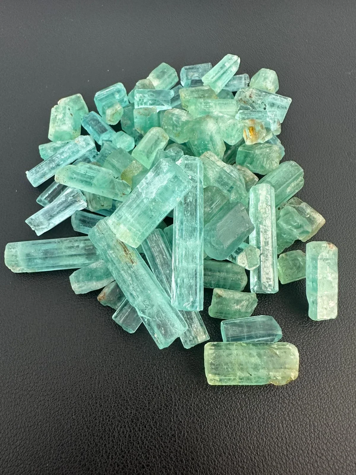 A pile of rough emerald crystals from Nigeria, pale bluish green six sided prisms with fine lines along their length, on a black leather surface