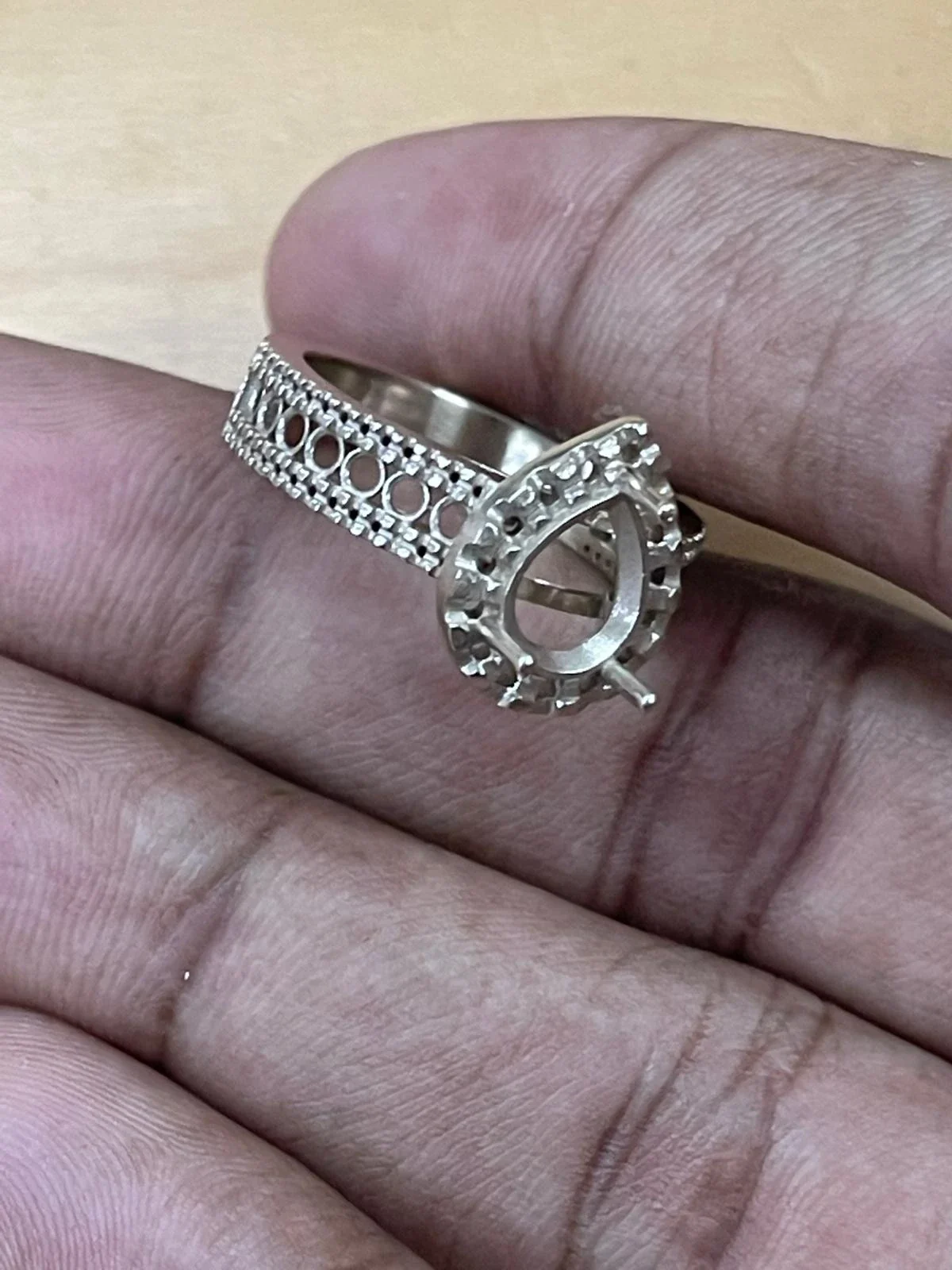 A bare white gold pear shaped halo ring mount held between two fingers, with empty seats and prongs waiting for stones