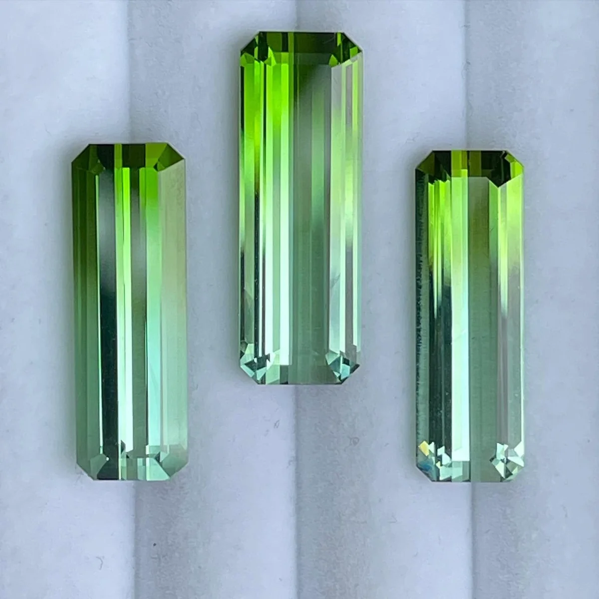 Three long emerald cut green tourmalines standing upright, each shading from lime green at the top to cool blue green at the base