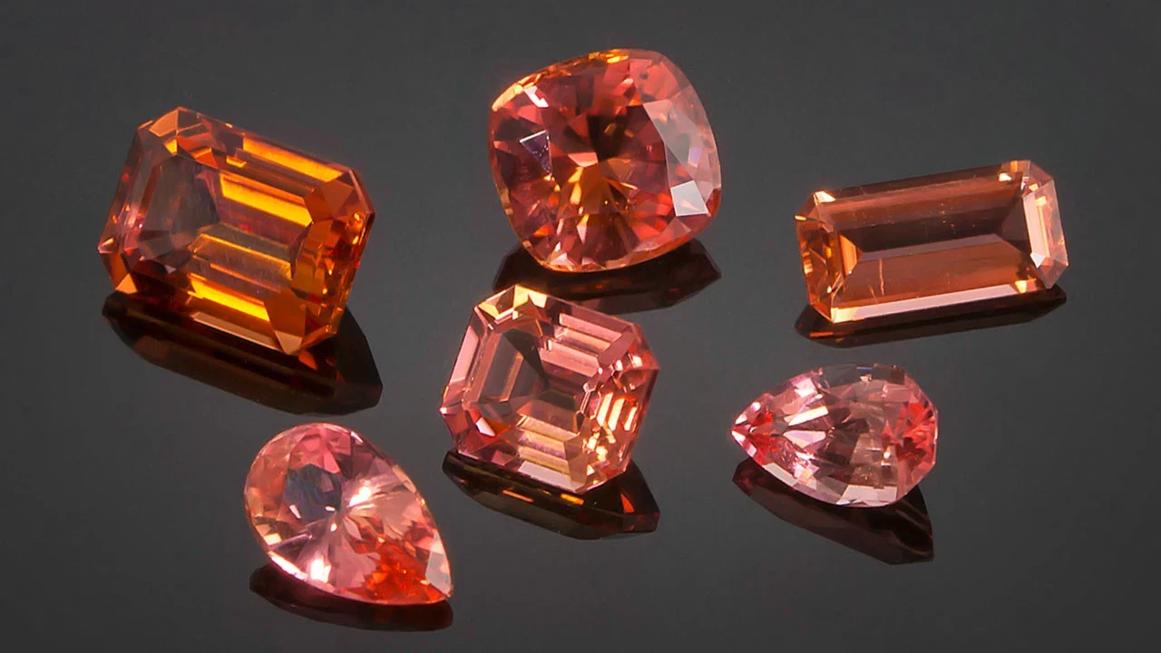 Six faceted orange, salmon and pink gemstones in emerald, cushion, pear and step cuts on a dark reflective surface