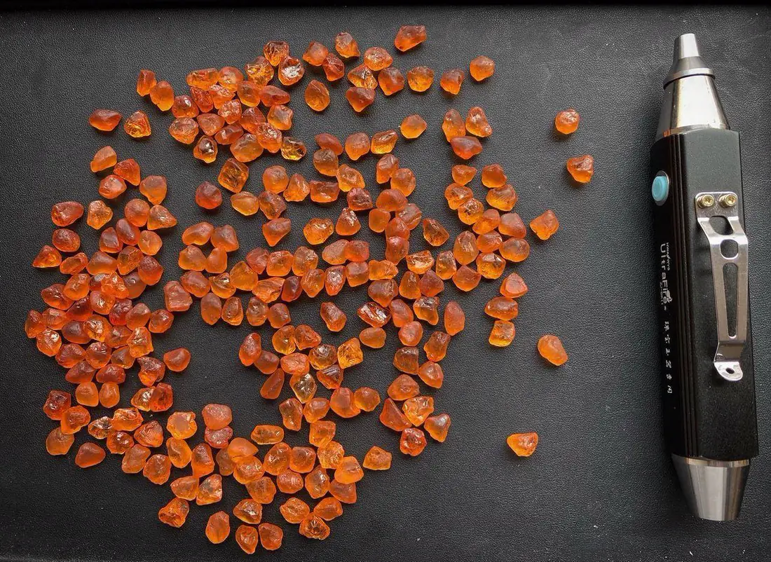A large parcel of orange spessartite garnet rough spread across a dark surface beside a gemmological torch